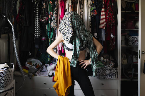 Dresses covering woman's face while standing with hands on hip against clothes rack in wardrobe - JCMF01760