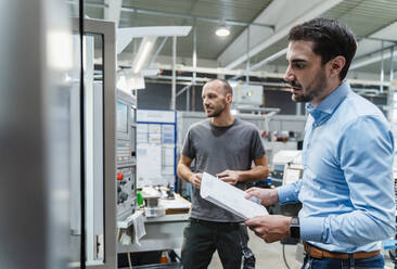 Male engineer with document examining machinery while standing with maintenance worker in background at factory - DIGF13729