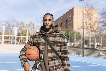 Junger Mann mit cooler Einstellung hält Basketball im Sportplatz an einem sonnigen Tag - JCCMF00316