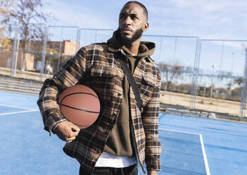 Young man with basketball looking away while standing in sports court on sunny day - JCCMF00317