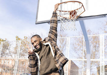 Glücklicher junger Mann, der Frieden gestikuliert, während er an einem sonnigen Tag am Basketballkorb hängt - JCCMF00323