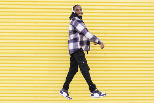 Cheerful young man jumping while listening music through headphones against yellow corrugated iron - JCCMF00332