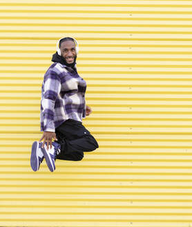 Happy male rapper listening music while jumping against yellow corrugated wall - JCCMF00333