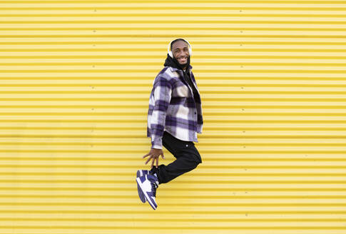 Happy young man in mid-air listening music through headphones against yellow corrugated iron - JCCMF00334