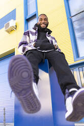 Fröhlicher männlicher Rapper mit den Händen in den Taschen, der die Schuhsohle gegen ein Gebäude zeigt - JCCMF00353