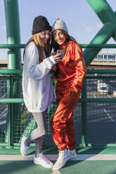 Smiling female friends using mobile phone while standing on bridge - JRVF00025