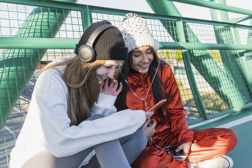 Lächelnde Freunde mit Kopfhörern und Handy auf einer Brücke sitzend - JRVF00029