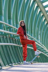 Junge Frau steht mit Skateboard und lehnt sich an ein Brückengeländer - JRVF00031