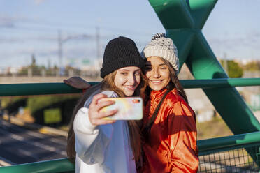 Smiling friends taking selfie through mobile phone while standing on bridge - JRVF00042