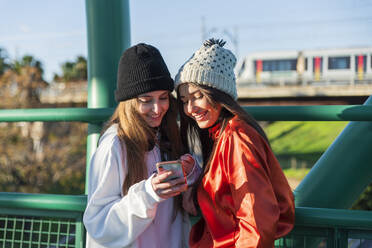 Lächelnde Freunde mit Mobiltelefon auf einer Brücke stehend - JRVF00043