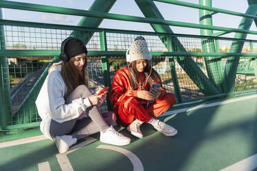 Female friends wearing knit hat and headphones using mobile phones while sitting on bridge - JRVF00044