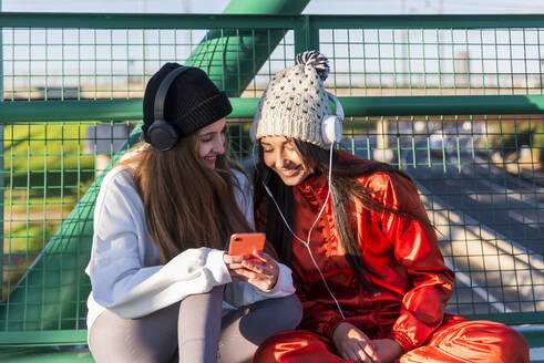 Freundinnen lächelnd bei der Nutzung eines Smartphones auf einer Brücke sitzend - JRVF00046