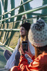 Frau mit Strickmütze, die auf einer Brücke sitzend einen lächelnden Freund mit dem Handy fotografiert - JRVF00048