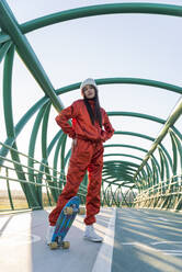 Junge Frau mit Händen in den Taschen steht mit Skateboard auf einer Brücke - JRVF00050