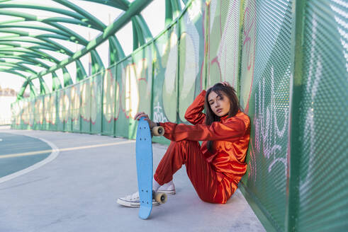 Junge Frau hält Skateboard und sitzt mit der Hand im Haar auf einer Brücke - JRVF00053