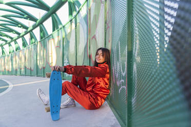 Smiling woman with skateboard resting while sitting on bridge - JRVF00054