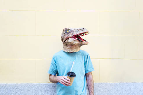 Mittlerer erwachsener Mann mit Dinosauriermaske und Kaffee an einer Wand in der Stadt stehend - JCMF01769