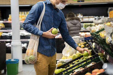 Midsection of man wearing face mask buying granny smith apples in supermarket - AFVF07952