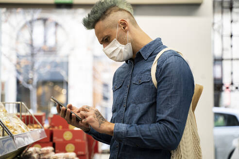Stilvoller Mann mit Gesichtsmaske, der im Supermarkt ein Smartphone benutzt - AFVF07954