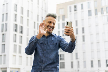 Glücklicher Mann, der während eines Videoanrufs gegen ein Gebäude mit seinem Smartphone winkt - AFVF07967