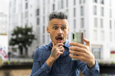 Schockierter Mann hält Smartphone während Videoanruf gegen Gebäude - AFVF07968