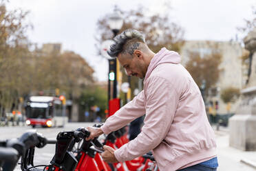 Stylish man renting bicycle through smart phone at parking station - AFVF07983