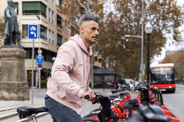 Stylish man looking away while renting bicycle at parking station - AFVF07984