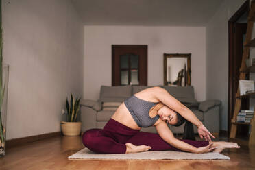 Young woman with arms raised practicing yoga while sitting on mat at home - GRCF00593