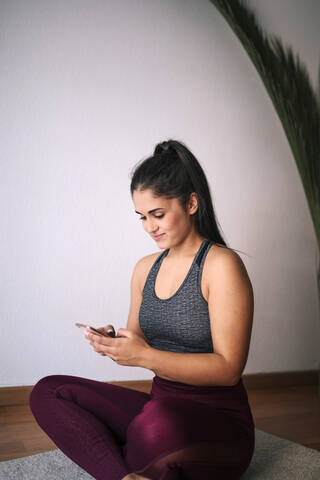 Frau benutzt ihr Smartphone, während sie zu Hause auf einer Übungsmatte an der Wand sitzt, lizenzfreies Stockfoto