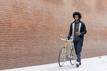 Junger Mann mit Kaffee in der Hand, der mit dem Fahrrad auf der Straße an einer Mauer entlang fährt - OCMF01938