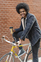 Lächelnder junger Mann mit Kaffee in der Hand, auf einem Fahrrad sitzend, an einer Backsteinmauer - OCMF01939