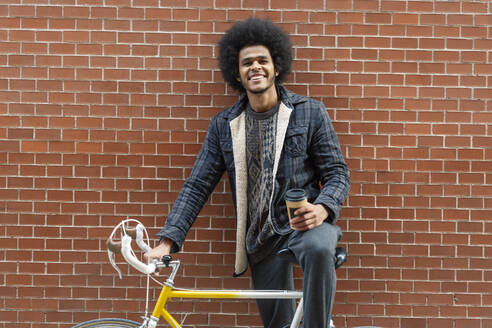 Young man holding coffee while standing with bicycle against brick wall - OCMF01940