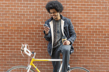 Junger Mann hält Kaffee und benutzt sein Smartphone, während er mit dem Fahrrad an einer Mauer steht - OCMF01941