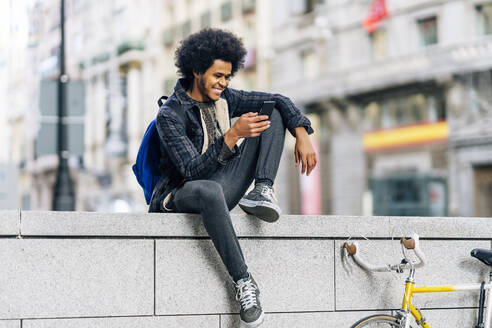 Lächelnder Mann mit lockigem Haar, der ein Mobiltelefon benutzt, während er auf einer Stützmauer in der Stadt sitzt - OCMF01947