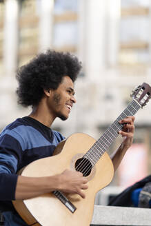 Junger Mann mit lockigem Haar, der im Freien stehend Gitarre spielt - OCMF01948