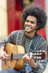 Lächelnder junger Mann mit lockigem Haar, der im Freien sitzend Gitarre spielt - OCMF01954