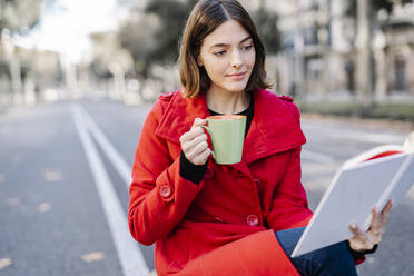 Junge Frau in Winterjacke trinkt Kaffee und liest ein Buch auf der Straße - JCZF00373