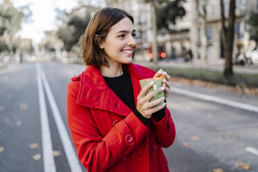 Smiling young woman holding coffee cup while looking away on street - JCZF00387