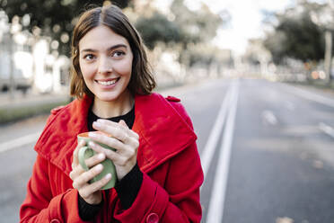 Smiling woman in winter jacket day dreaming while holding coffee cup on road - JCZF00388