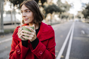 Nachdenkliche junge Frau in roter Jacke mit Kaffeetasse auf der Straße - JCZF00393