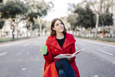 Young woman in red jacket with coffee cup day dreaming while holding book on road - JCZF00398