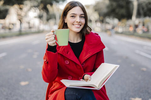 Smiling young woman with book having coffee while sitting on seat at street stock photo
