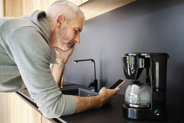 Mature man with hand on chin using mobile phone while leaning on kitchen counter at home - AODF00134