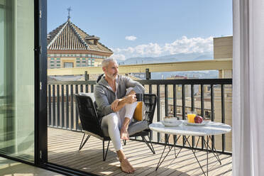 Nachdenklicher reifer Mann sitzt auf einem Stuhl mit Frühstück auf dem Balkon an einem sonnigen Morgen - AODF00136