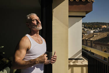 Mature man with eyes closed holding coffee cup while standing in sunlight - AODF00139