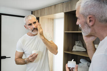 Mature man applying face cream while looking in mirror at bathroom - AODF00147
