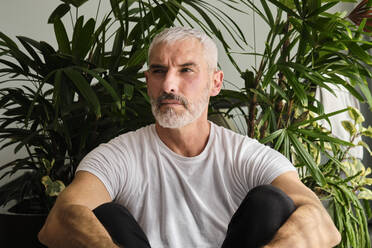 Handsome mature man contemplating while sitting against plant at home - AODF00157