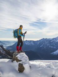 Männlicher Wanderer, der sein Smartphone benutzt, während er auf einem verschneiten Berg gegen den Himmel steht, Orobische Alpen, Lecco, Italien - MCVF00695