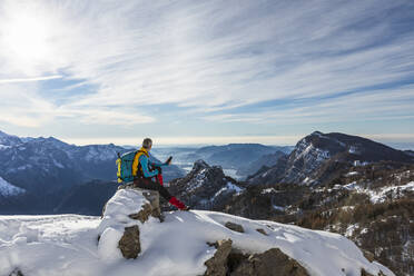 Männlicher Wanderer, der sein Handy benutzt, während er auf einem Berg gegen den Himmel sitzt, Orobic-Alpen, Lecco, Italien, im Winter - MCVF00697