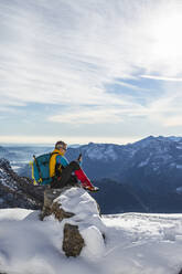 Älterer Mann mit Rucksack, der ein Mobiltelefon benutzt, während er auf einem verschneiten Berg sitzt, Orobic-Alpen, Lecco, Italien - MCVF00698